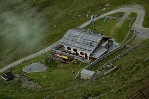 A rustic building with a wooden roof is nestled within a grassy landscape. It is surrounded by a fence and situated near a gravel path that curves alongside the property. There are small animal enclosures visible and a few animals grazing nearby. Mist lightly covers parts of the green hillside, adding to the serene atmosphere.