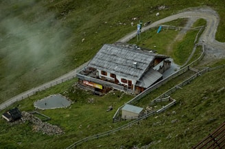 A rustic building with a wooden roof is nestled within a grassy landscape. It is surrounded by a fence and situated near a gravel path that curves alongside the property. There are small animal enclosures visible and a few animals grazing nearby. Mist lightly covers parts of the green hillside, adding to the serene atmosphere.