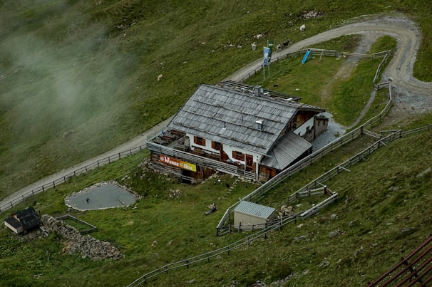 A rustic building with a wooden roof is nestled within a grassy landscape. It is surrounded by a fence and situated near a gravel path that curves alongside the property. There are small animal enclosures visible and a few animals grazing nearby. Mist lightly covers parts of the green hillside, adding to the serene atmosphere.