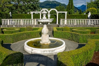 A beautifully manicured garden features a central stone fountain surrounded by neatly trimmed hedges arranged in intricate patterns. The garden is flanked by white balustrades and statues, set against a backdrop of lush green trees and distant mountains.