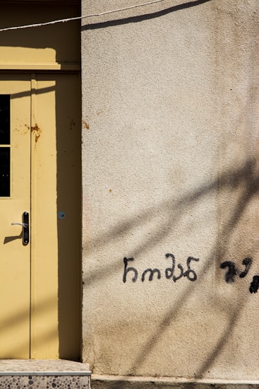 A textured wall with a light yellow door on the left, featuring a metallic door handle and visible rust marks. Graffiti in a foreign script is painted in black on the beige wall surface, accompanied by shadows of wires or cables crossing the wall.