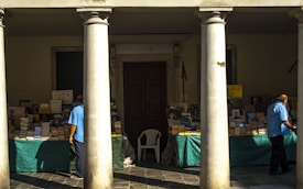 An outdoor bookstall is set up under a covered area, flanked by large columns. The tables are covered with green cloths beneath stacks of books, some of which are marked with signs indicating discounts. Two men, wearing blue shirts, are browsing the selection. The scene is bathed in afternoon sunlight, casting strong shadows across the pavement.