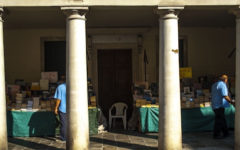 An outdoor bookstall is set up under a covered area, flanked by large columns. The tables are covered with green cloths beneath stacks of books, some of which are marked with signs indicating discounts. Two men, wearing blue shirts, are browsing the selection. The scene is bathed in afternoon sunlight, casting strong shadows across the pavement.