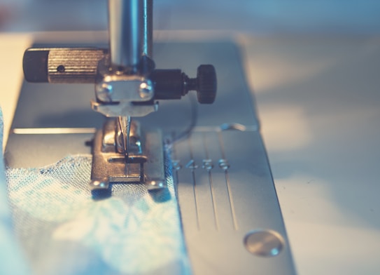 A close-up view of a sewing machine needle and presser foot, stitching fabric. The image focuses on the metallic parts of the machine and the blue fabric being sewn, with soft lighting creating a serene ambiance.