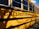 A school bus is parked with the side displaying the words "Chelmsford Public Schools" in bold black letters. The yellow exterior and large windows are visible, casting shadows from nearby trees or surroundings.