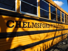 A school bus is parked with the side displaying the words "Chelmsford Public Schools" in bold black letters. The yellow exterior and large windows are visible, casting shadows from nearby trees or surroundings.