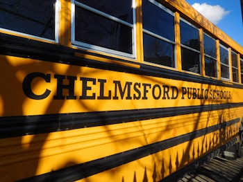A school bus is parked with the side displaying the words "Chelmsford Public Schools" in bold black letters. The yellow exterior and large windows are visible, casting shadows from nearby trees or surroundings.