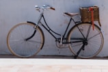 A vintage-style bicycle with a brown leather seat and handlebars leans against a white wall. It features a classic design with thin tires and a basket attached to the rear with a red ribbon hanging from it.
