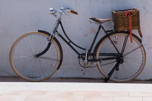A vintage-style bicycle with a brown leather seat and handlebars leans against a white wall. It features a classic design with thin tires and a basket attached to the rear with a red ribbon hanging from it.