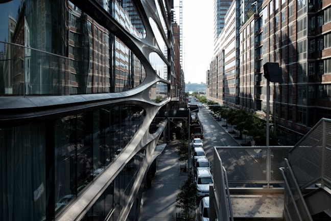 A modern urban landscape with high-rise buildings lining a narrow street. The architecture features futuristic design elements with reflective glass surfaces. Several vehicles are parked along the road, and trees provide some greenery along the sidewalk. The lighting suggests early morning or late afternoon, casting long shadows.