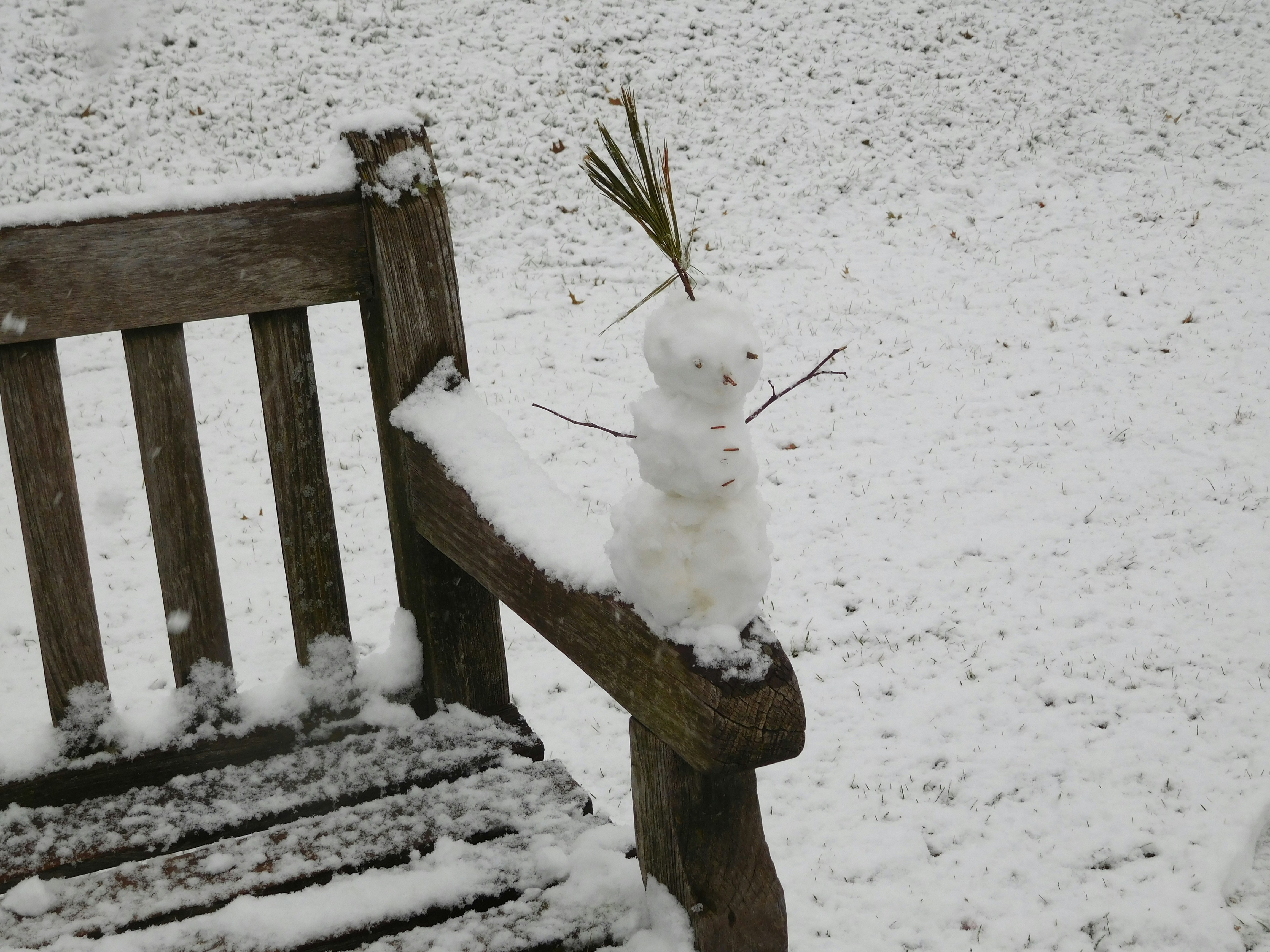 Snow-covered scene featuring a tiny snowman perched on a frosted wooden railing beside a weathered bench.