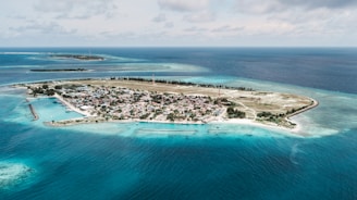 A small island surrounded by clear blue ocean waters, featuring a developed area with buildings, possibly a town or village, in the center. There are boats visible near the shoreline, and the island is bordered by patches of greenery and sandy beaches. The horizon showcases more small land masses in the distance beneath a partly cloudy sky.