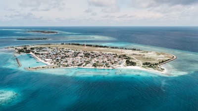 A small island surrounded by clear blue ocean waters, featuring a developed area with buildings, possibly a town or village, in the center. There are boats visible near the shoreline, and the island is bordered by patches of greenery and sandy beaches. The horizon showcases more small land masses in the distance beneath a partly cloudy sky.