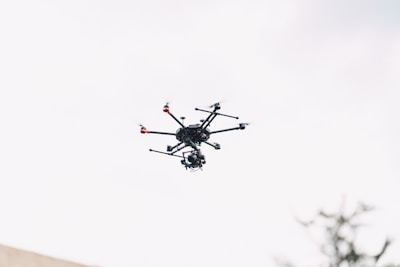 A drone with multiple propellers is flying in a clear sky. The drone has a complex frame with visible landing gear and sensors attached. The sky is bright, providing high contrast to the dark color of the drone.