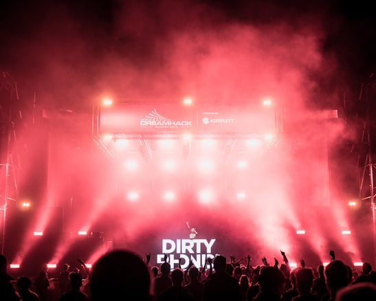 A vibrant concert scene with a large crowd silhouetted against bright stage lights. The atmosphere is filled with smoke, enhancing the dramatic lighting effects. A prominent banner displays the event name 'Dreamhack Summer 2019,' and a DJ booth with the name 'Dirtyphonics' is visible.