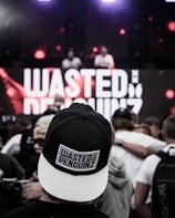 A crowd at a live music event, focused on a person wearing a black and white cap with the text 'WASTED PENGUINZ' on it. In the background, a large screen displays the same text, with two DJs performing.
