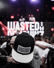 A crowd at a live music event, focused on a person wearing a black and white cap with the text 'WASTED PENGUINZ' on it. In the background, a large screen displays the same text, with two DJs performing.
