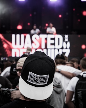A crowd at a live music event, focused on a person wearing a black and white cap with the text 'WASTED PENGUINZ' on it. In the background, a large screen displays the same text, with two DJs performing.