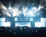 A vibrant concert scene with dynamic lighting effects featuring bright beams and illuminated screens. The audience, silhouetted against the lights, captures the event on their smartphones.