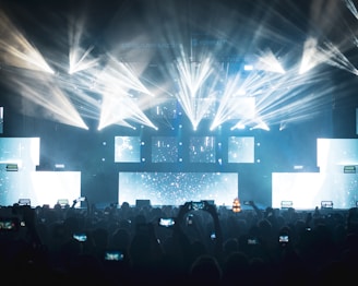 A vibrant concert scene with dynamic lighting effects featuring bright beams and illuminated screens. The audience, silhouetted against the lights, captures the event on their smartphones.