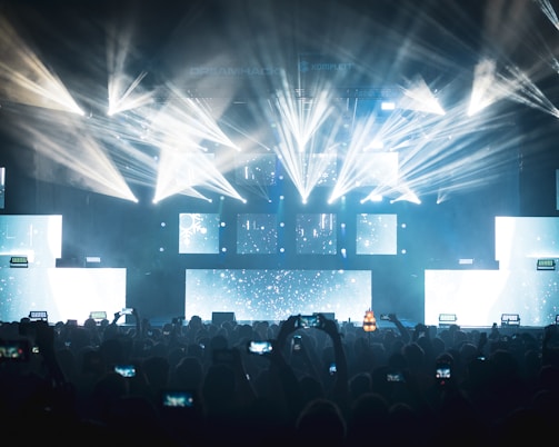 A vibrant concert scene with dynamic lighting effects featuring bright beams and illuminated screens. The audience, silhouetted against the lights, captures the event on their smartphones.
