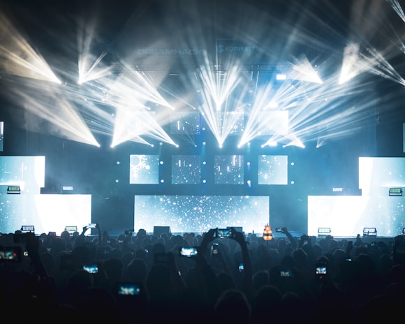 A vibrant concert scene with dynamic lighting effects featuring bright beams and illuminated screens. The audience, silhouetted against the lights, captures the event on their smartphones.