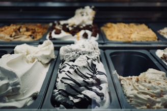 A variety of gelato flavors are arranged in metal containers at a gelato shop. The selection includes creamy white flavors with chocolate drizzle and various nut toppings.