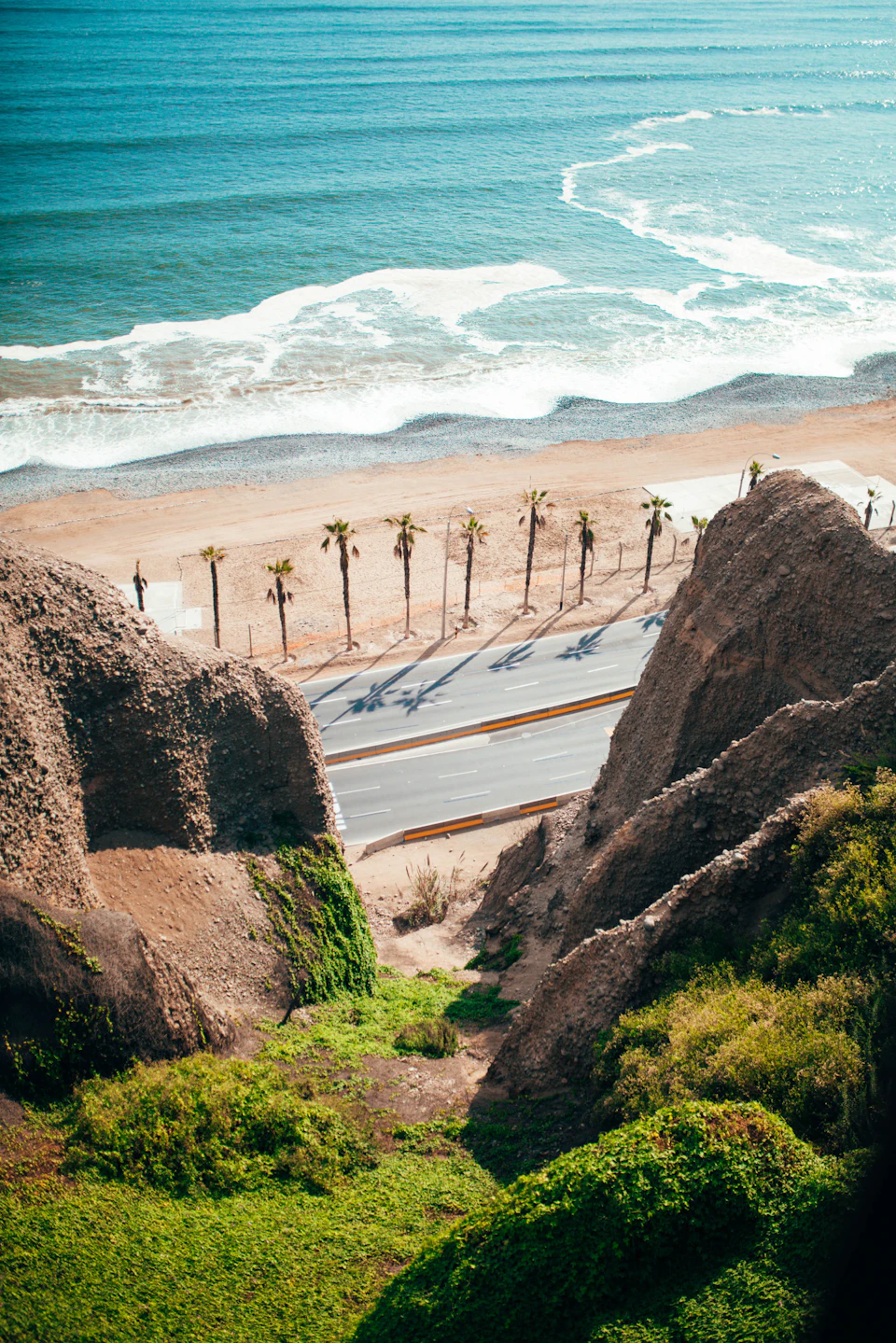 The image presents an aerial view of a beachfront road, with palm trees lining both sides and a body of water visible on the left side. The road is marked by a white line running down its center, dividing it into two lanes. On the right side of the road, there are several palm trees standing tall against the sky.

The sky above the beach is a clear blue color with some clouds scattered across it. The water below mirrors this blue hue and appears to be calm, reflecting the serene atmosphere of the scene.