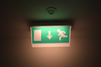 An illuminated emergency exit sign is mounted on the ceiling, featuring a green background with white symbols indicating a direction to an exit. The symbols include a door, a downward arrow, and a running figure.
