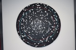 A circular abstract artwork with a galaxy-like appearance in a dark color palette. It features swirling patterns with white splatters and lines, creating a sense of movement. Red and blue accents punctuate the predominantly black background.
