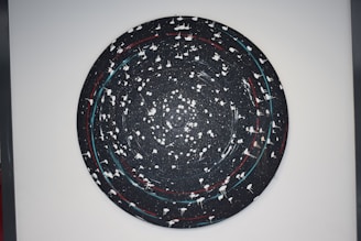 A circular abstract artwork with a galaxy-like appearance in a dark color palette. It features swirling patterns with white splatters and lines, creating a sense of movement. Red and blue accents punctuate the predominantly black background.