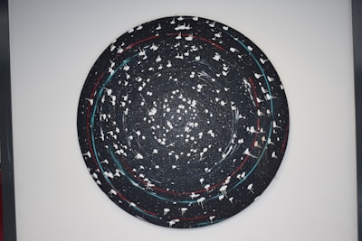 A circular abstract artwork with a galaxy-like appearance in a dark color palette. It features swirling patterns with white splatters and lines, creating a sense of movement. Red and blue accents punctuate the predominantly black background.