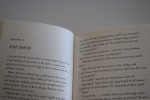 An open book displaying a double-page spread with text. The left page starts with a date, September 18, followed by the address 'DEAR MARTIN,'. The text appears to be part of a letter or a diary entry, discussing personal thoughts and experiences related to school and a person named Mama. The right page continues this narrative, mentioning topics such as a cop, handcuffs, and a classroom discussion. The book is held at an angle, with lighting casting a mild shadow over the pages.