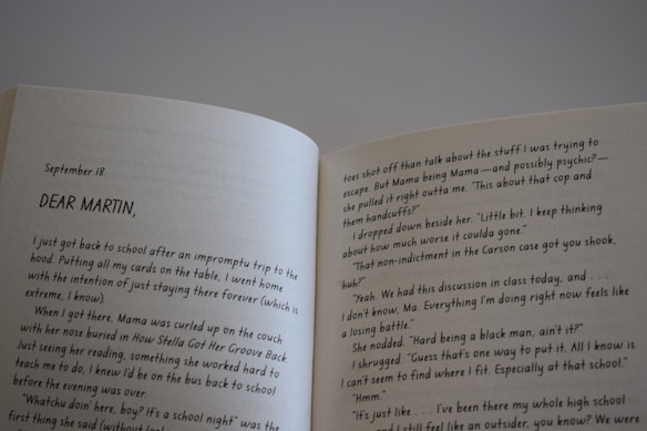 An open book displaying a double-page spread with text. The left page starts with a date, September 18, followed by the address 'DEAR MARTIN,'. The text appears to be part of a letter or a diary entry, discussing personal thoughts and experiences related to school and a person named Mama. The right page continues this narrative, mentioning topics such as a cop, handcuffs, and a classroom discussion. The book is held at an angle, with lighting casting a mild shadow over the pages.