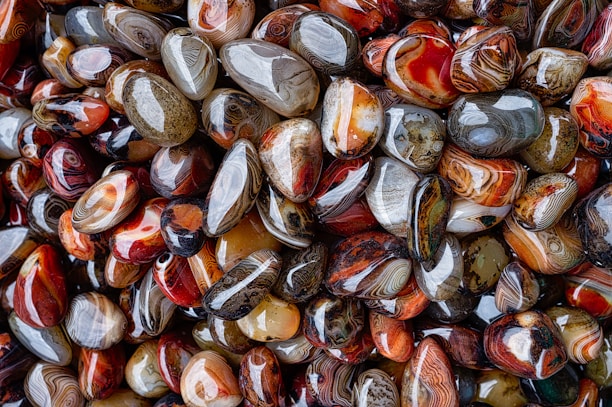 An assortment of polished and colorful agate stones, displaying a range of intricate patterns and swirls. The stones vary in color, with shades of red, brown, gray, and orange prominently featured. Their smooth, glossy surfaces reflect light, enhancing their vibrant appearance.