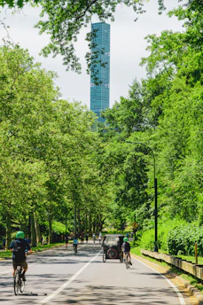 A vibrant scene of tourists enjoying bike rides on tree-lined paths just steps from Central Park, with the NYC skyline in the background.
