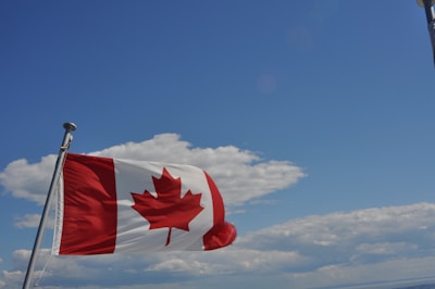 A Canadian flag waves prominently against a backdrop of blue sky and fluffy clouds. The red maple leaf on the flag stands out vividly, contrasting with the white and red sections of the flag.