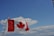 A Canadian flag waves prominently against a backdrop of blue sky and fluffy clouds. The red maple leaf on the flag stands out vividly, contrasting with the white and red sections of the flag.