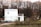 A white, two-story modular building is situated in a rural area with a backdrop of leafless trees and overgrown grass. The building has a simple design with small windows and a fenced perimeter. There is a cartoon character figure near the right side of the building.