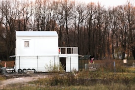 A white, two-story modular building is situated in a rural area with a backdrop of leafless trees and overgrown grass. The building has a simple design with small windows and a fenced perimeter. There is a cartoon character figure near the right side of the building.