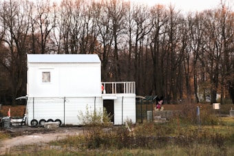 A white, two-story modular building is situated in a rural area with a backdrop of leafless trees and overgrown grass. The building has a simple design with small windows and a fenced perimeter. There is a cartoon character figure near the right side of the building.