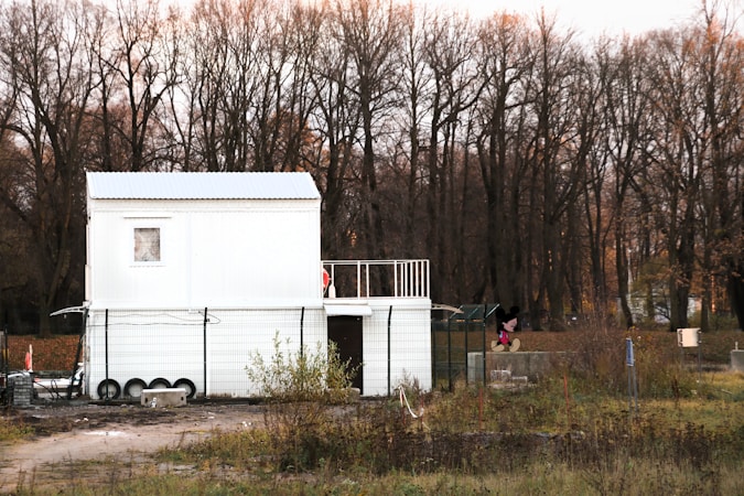 A white, two-story modular building is situated in a rural area with a backdrop of leafless trees and overgrown grass. The building has a simple design with small windows and a fenced perimeter. There is a cartoon character figure near the right side of the building.