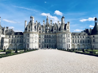 A grand Renaissance-style chateau with intricate stonework and numerous spires and turrets. The building is set against a bright blue sky with a few clouds, surrounded by manicured gardens and a gravel courtyard.