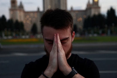 A person with closed eyes appears to be in a moment of meditation or prayer, with hands pressed together near the face in a gesture of contemplation. The background features a large, ornate building with soft lighting and blurred greenery.