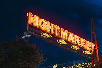 A neon-lit sign featuring the words 'Night Market' illuminated in bright orange and yellow hues stands prominently against a dark evening sky. The sign is elevated on a metal structure, with arrows pointing below.