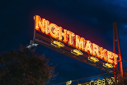A neon-lit sign featuring the words 'Night Market' illuminated in bright orange and yellow hues stands prominently against a dark evening sky. The sign is elevated on a metal structure, with arrows pointing below.