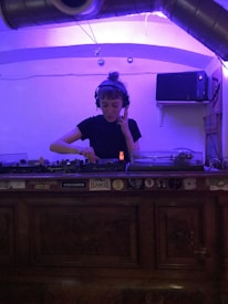 A person is wearing headphones and using DJ equipment in a dimly lit room. The equipment is placed on a wooden surface adorned with various stickers. The room is illuminated with purple lighting, and there are industrial ventilation pipes visible on the ceiling.
