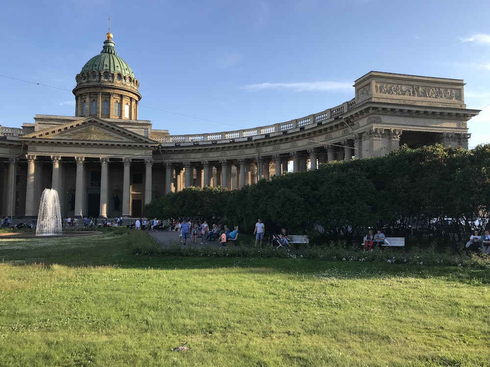 people standing and sitting on pathway beside trees, fountain, and building during day