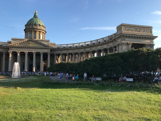 people standing and sitting on pathway beside trees, fountain, and building during day in Kazan Cathedral Russia