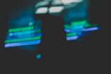 A blurred image featuring overlapping horizontal streaks of neon green and blue light against a dark background, creating an abstract and dynamic visual effect.
